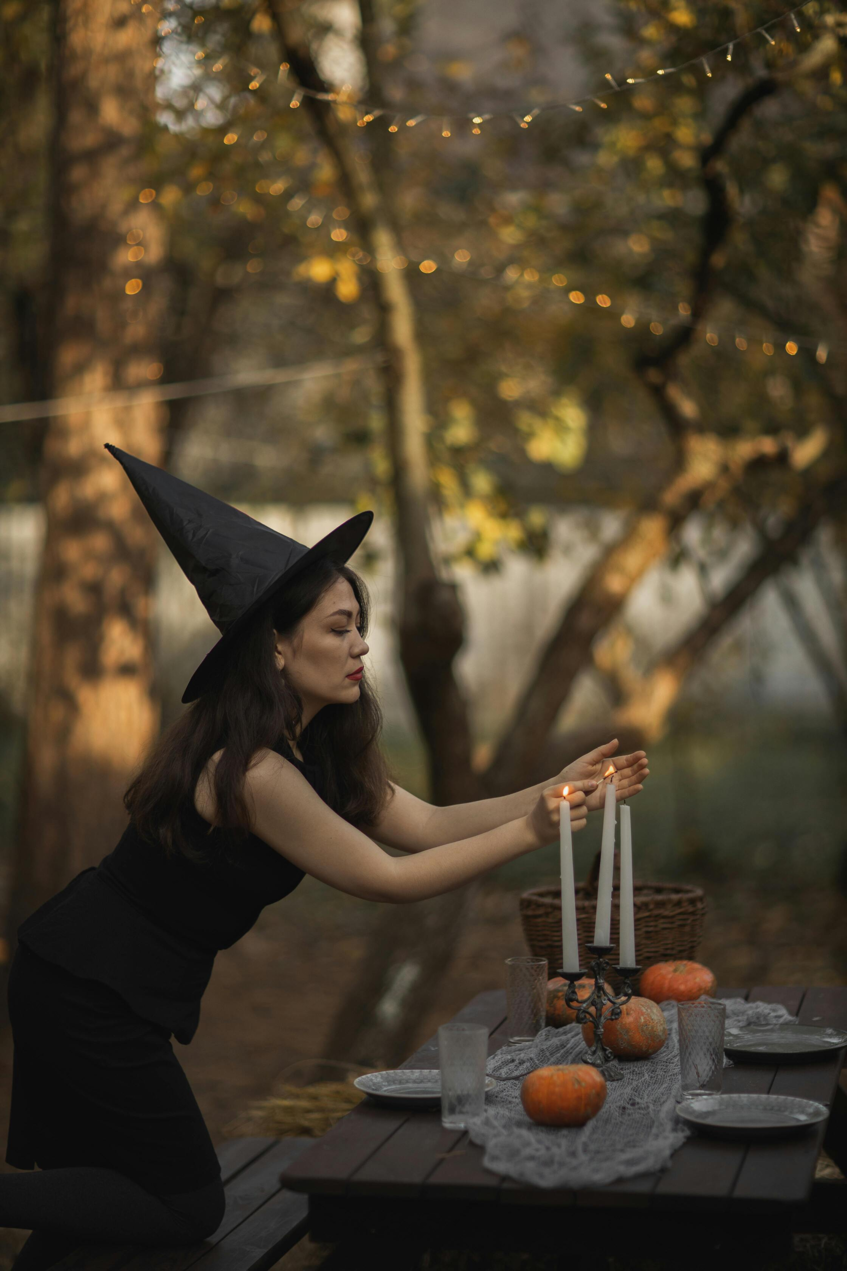 Woman in a black witch hat lighting tall white candles at a decorated outdoor table with pumpkins and fall leaves
